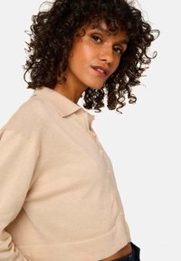 Pull beige cropped avec une texture douce, col relevé et fermeture à boutons, présentant des accents distinctifs sur les manches côtelées.