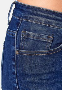 Blue denim jeans featuring a high-waisted design, five-pocket style, subtle fading, and contrast stitching. Back pocket detail visible.