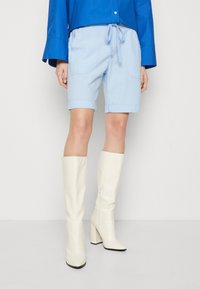 Los pantalones cortos casuales de color azul claro presentan dobladillos enrollados, una cintura con cordón y bolsillos laterales. Se combinan con botas de tacón cuadrado blancas hasta la rodilla.