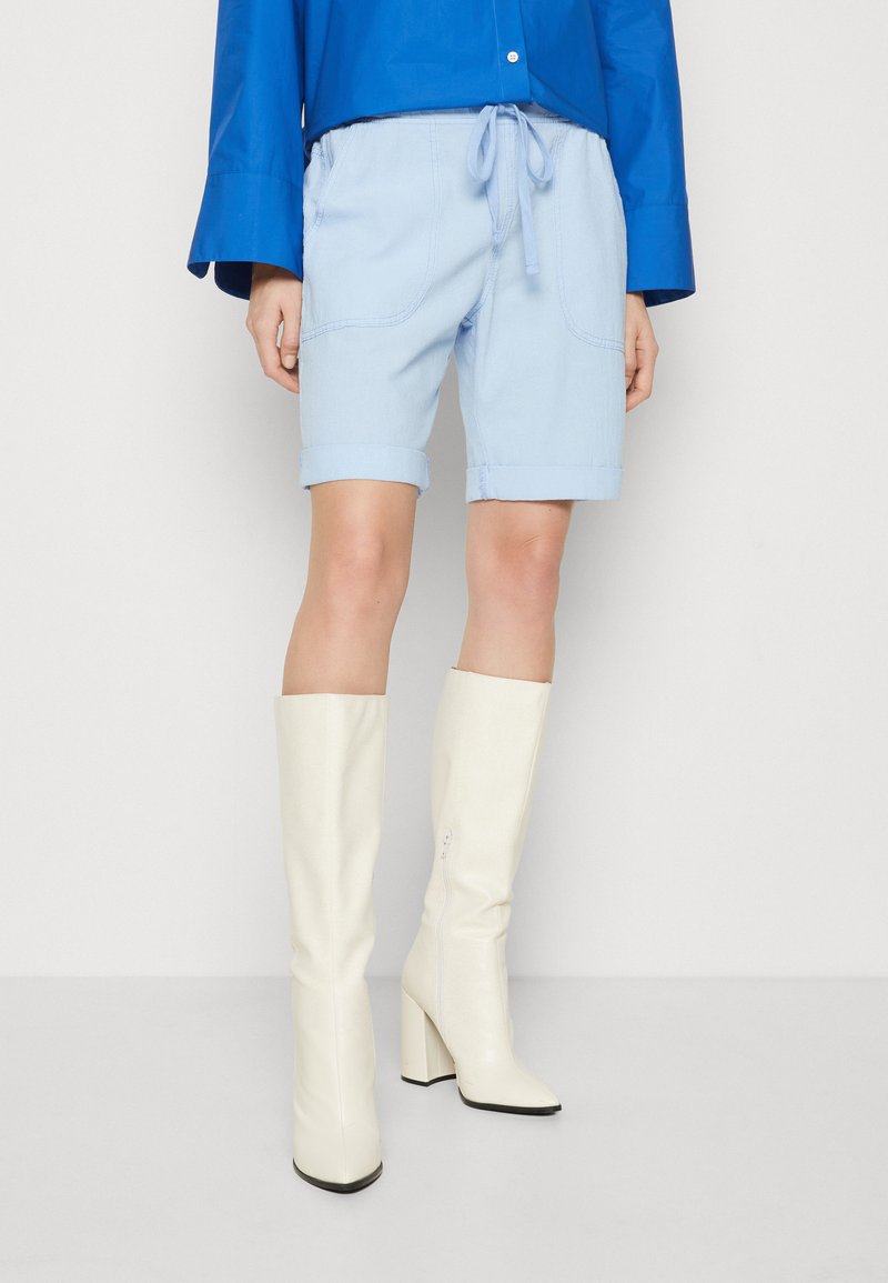 Los pantalones cortos casuales de color azul claro presentan dobladillos enrollados, una cintura con cordón y bolsillos laterales. Se combinan con botas de tacón cuadrado blancas hasta la rodilla.