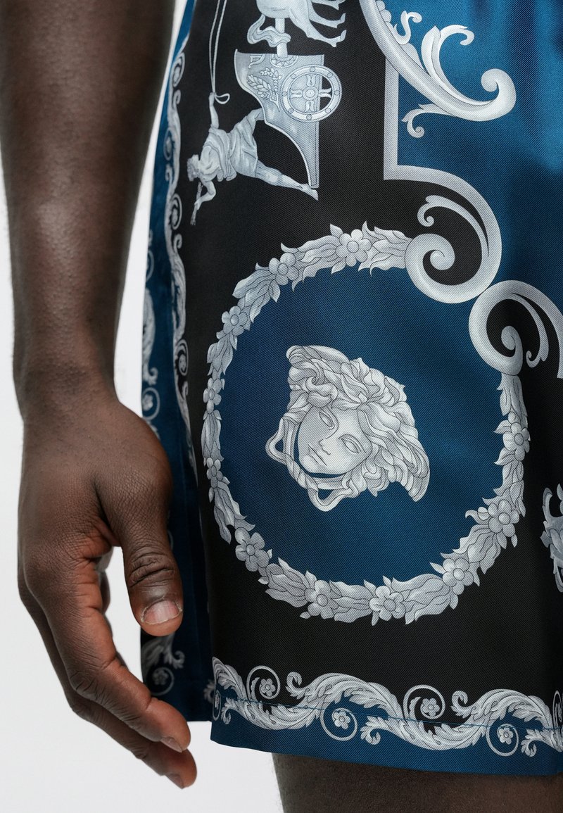 Blå og sorte shorts i glat stof med udsmykkede sølvmønstre, herunder blomsterdetaljer og et Medusa-lignende ansigtsdesign.