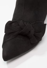 Chaussure en daim noir à bout pointu avec un grand nœud décoratif sur le dessus, photographiée sur fond blanc.