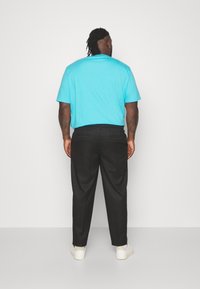 Man met tatoeages in een helderblauwe kraagloze shirt, zwarte broek en witte sneakers, staand met zijn gezicht naar een effen witte muur.
