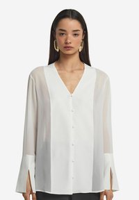 Blusa bianca a maniche lunghe con scollatura a V profonda, bottoni frontali e polsini con spacco; tessuto trasparente con una texture liscia; design minimalista.