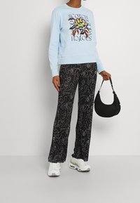 Ljusblå sweatshirt med grafisk design, svarta mönstrade vida byxor med abstrakta former och vita sneakers. Svart väska hålls i handen.