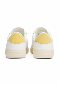 Baskets blanches avec un dessus en cuir lisse, un accent de talon en daim jaune, et une semelle en caoutchouc beige portant "TOMMY JEANS" embossé sur le côté.