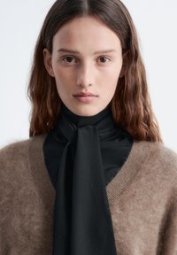 Brauner, flauschiger Pullover mit einer schwarzen Seidenkrawatte, die einen flachen Knoten aufweist. Die glatte Textur und das weiche Finish stehen im Kontrast zum Strickmuster des Pullovers.