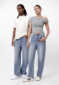 Lys grå cropped topp på en kvinne, løse blå jeans på begge, mørk skjorte på en mann. Sneakers i hvitt med svarte detaljer. Nøytral bakgrunn.