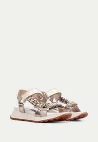 Kultaiset metalliset sandaalit, joissa on punotut hihnat, pehmeä pohja ja teksturoitu kumipohja. Säädettävät nilkka- ja jalkahihnat.