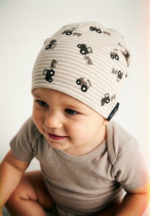 Lächelndes Kleinkind, das eine beigefarbene gestreifte Mütze mit Traktoraufdrucken und einen passenden beigefarbenen Kurzarmstrampler trägt und vor einem schlichten Hintergrund sitzt.