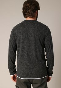 Man met zijn rug naar de camera, gekleed in een donkergrijze longsleeve shirt over een wit shirt en olijfgroene broek, tegen een effen achtergrond.