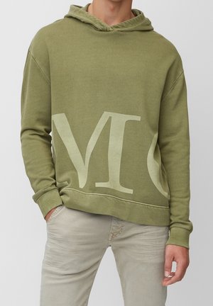 Mężczyzna w oliwkowo-zielonej bluzie z kapturem z dużymi, beżowymi częściowymi literami oraz jasnobeżowych spodniach, stojący z jedną ręką w kieszeni.