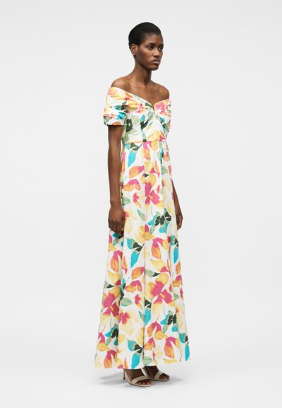 Γυναίκα με πολύχρωμο μάξι φόρεμα με φλοράλ σχέδια και ακάλυπτους ώμους, με φουσκωτά μανίκια, που στέκεται πλαγίως μπροστά σε απλό λευκό φόντο.