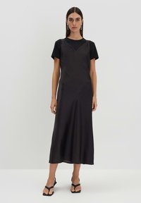 Schwarzes, knielanges Slipkleid mit V-Ausschnitt und strukturierter Oberfläche, über einem kurzärmeligen schwarzen T-Shirt getragen. Kombiniert mit schwarzen Sandalen.