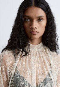 Spitzenbluse mit hohem Ausschnitt und gefältelten Details; durchscheinendes florales Muster in Cremefarbe über einer dunklen Unterschicht.