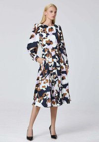 Femme se tenant avec une main dans la poche, portant une robe midi à manches longues avec des motifs abstraits marron, noir, blanc et lavande, et des talons noirs.