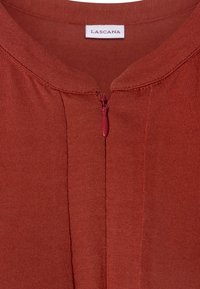 Bordeauxfärgad topp med rund halsringning, framzip, och ribbat mönster, med en lapp märkt "Lascana" på nacken.