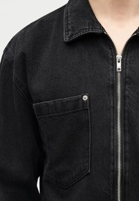 Μαύρο τζιν μπουφάν με μπροστινή τσέπη στο στήθος που ασφαλίζει με μεταλλικό καρφί και κλείσιμο με ασημένιο φερμουάρ.