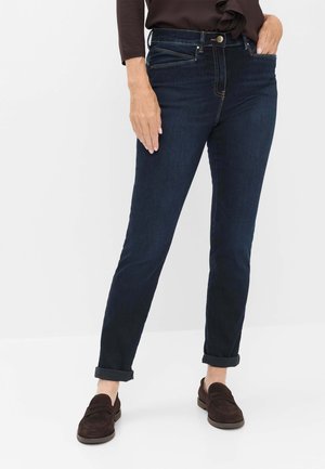 Person iført mørkeblå opsmøgede jeans og brune loafers, med den ene hånd i lommen og den anden hvilende langs benet.