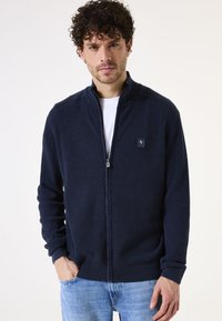 Sudadera de cremallera en color navy con tejido texturizado, puños y dobladillo acanalados, y un pequeño parche con logo en el pecho. Lleva puesta sobre una camiseta blanca.