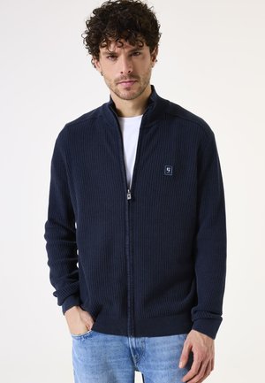 Sudadera de cremallera en color navy con tejido texturizado, puños y dobladillo acanalados, y un pequeño parche con logo en el pecho. Lleva puesta sobre una camiseta blanca.