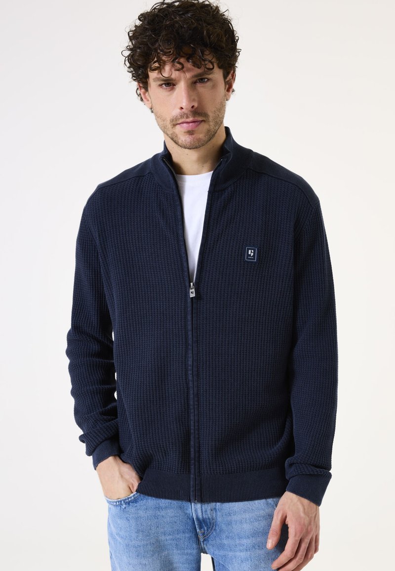 Sudadera de cremallera en color navy con tejido texturizado, puños y dobladillo acanalados, y un pequeño parche con logo en el pecho. Lleva puesta sobre una camiseta blanca.