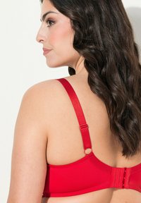 Red bra with adjustable straps and a hook-and-eye closure. Smooth fabric and rounded cups. Features a circular hardware accent on strap.