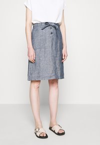 Woman wearing a knee-length blue chambray skirt with front buttons and tie belt, paired with a white top and beige sandals with round buckles.