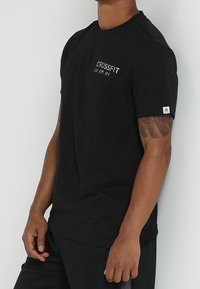 Černé bavlněné tričko s krátkým rukávem, s bílým logem "CROSSFIT" na hrudi a logem na rukávu. Hladká textura.