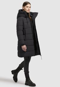 Abrigo de plumas negro, hasta las rodillas, diseño acolchado, capucha oversized, cremallera frontal, bolsillos laterales, combinado con pantalones ajustados negros y botas de tobillo.