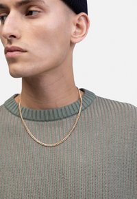 Grüner Strickpullover mit geripptem Muster, dazu eine goldene Kettenhalskette. Der Hintergrund ist neutral und lenkt die Aufmerksamkeit auf das Kleidungsstück und die Accessoires.