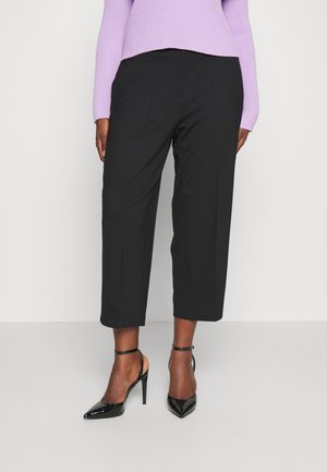 Frau trägt schwarze, verkürzte Hosen mit weitem Bein, schwarze hochhackige Schuhe mit Knöchelriemen und einen fliederfarbenen, gerippten Pullover.