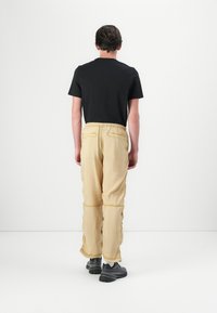 Mann står med ryggen til kameraet, iført svart t-skjorte, beige cargo bukser og svarte sko mot en enkel hvit bakgrunn.