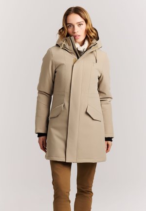 Vrouw die een beige winterjas met capuchon en steekzakken draagt en een bruine broek, staand tegen een egale lichte achtergrond.