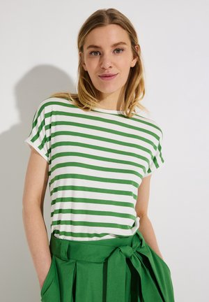 Junge Frau, die ein weiß-grün gestreiftes kurzärmeliges Oberteil und einen hoch taillierten grünen Rock mit gebundenem Gürtel trägt, vor einem schlichten Hintergrund stehend.