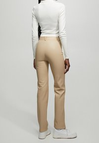 Pantalon beige sur mesure avec une coupe slim, taille haute et poches avant, associé à des baskets blanches et un haut blanc ajusté.