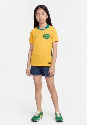 Jeune personne debout portant un maillot de football australien jaune, un short bleu foncé, et des baskets Nike vertes et jaunes sur fond blanc.