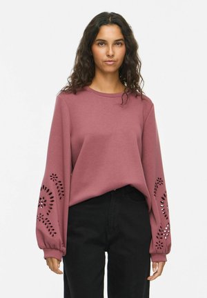 Femme portant un haut rose à manches longues avec des motifs floraux découpés sur les manches et un pantalon noir taille haute.