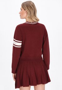 Burgundy neulemekko, jossa on pitkät hihat, ribba-helmarit ja pleated-hame. Hihansuissa on valkoiset raidalliset yksityiskohdat.