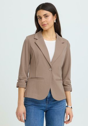 Vrouw draagt taupekleurige blazer met één knoop en geplooide mouwen over een wit topje en blauwe spijkerbroek, poseert tegen een effen lichte achtergrond.