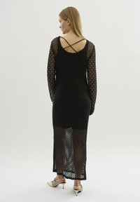 Rochie neagră din tricot cu mâneci lungi, având o textură mesh deschisă și bretele în cruce pe spate. Design până la gleznă, cu o siluetă ajustată.