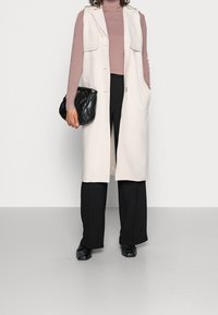 Lange crèmekleurige mouwloze vest over een roze coltrui, gecombineerd met zwarte broek met wijde pijpen en een zwarte textuurhandtas. Zwarte schoenen maken de outfit compleet.