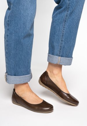 Feet wearing brown leather ballet flats paired with cuffed blue denim jeans against a plain background.
