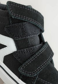 Svart och grå sneakers med en texturerad mockaovandel, vita accenter och kardborreband. Funktioner inkluderar sömnadsdetaljer och en gummisula.