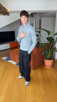 Γκρι πουλόβερ με πλεκτό σχέδιο ζακέτας και φερμουάρ, μαύρο παντελόνι άνετης εφαρμογής και ασημένια παπούτσια. Εσωτερικός χώρος με μεγάλο πράσινο φυτό σε γλάστρα.