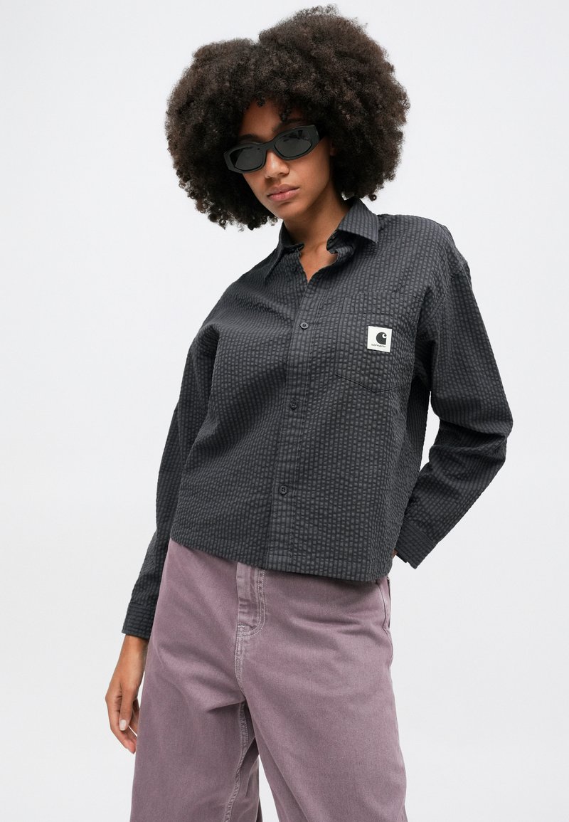 Schwarzes, strukturiertes Hemd mit Knöpfen und einem kleinen Logopatch, kombiniert mit helllila Hosen und dunklen rechteckigen Sonnenbrillen.