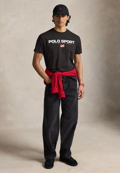 Νέος άνδρας που φοράει μαύρο t-shirt Polo Sport, μαύρο καπέλο, φαρδιά μαύρα τζιν, μαύρα παπούτσια, με κόκκινο πουλόβερ δεμένο γύρω από τη μέση.