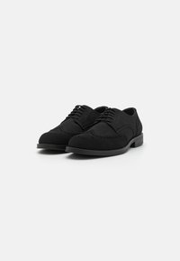 Schwarze Wildleder-Halbschuhe mit runder Zehenform, Schnürdesign, Broguemuster und einer flachen schwarzen Gummisohle.