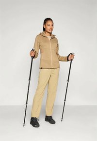 Giovane donna in piedi con bastoni da trekking neri, che indossa una giacca beige, pantaloni beige chiaro e scarponi da trekking neri, su uno sfondo neutro.