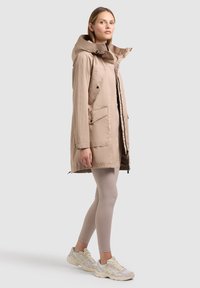Donna in piedi con parka beige con cappuccio, leggings beige chiaro e sneakers bianche su uno sfondo grigio chiaro uniforme.